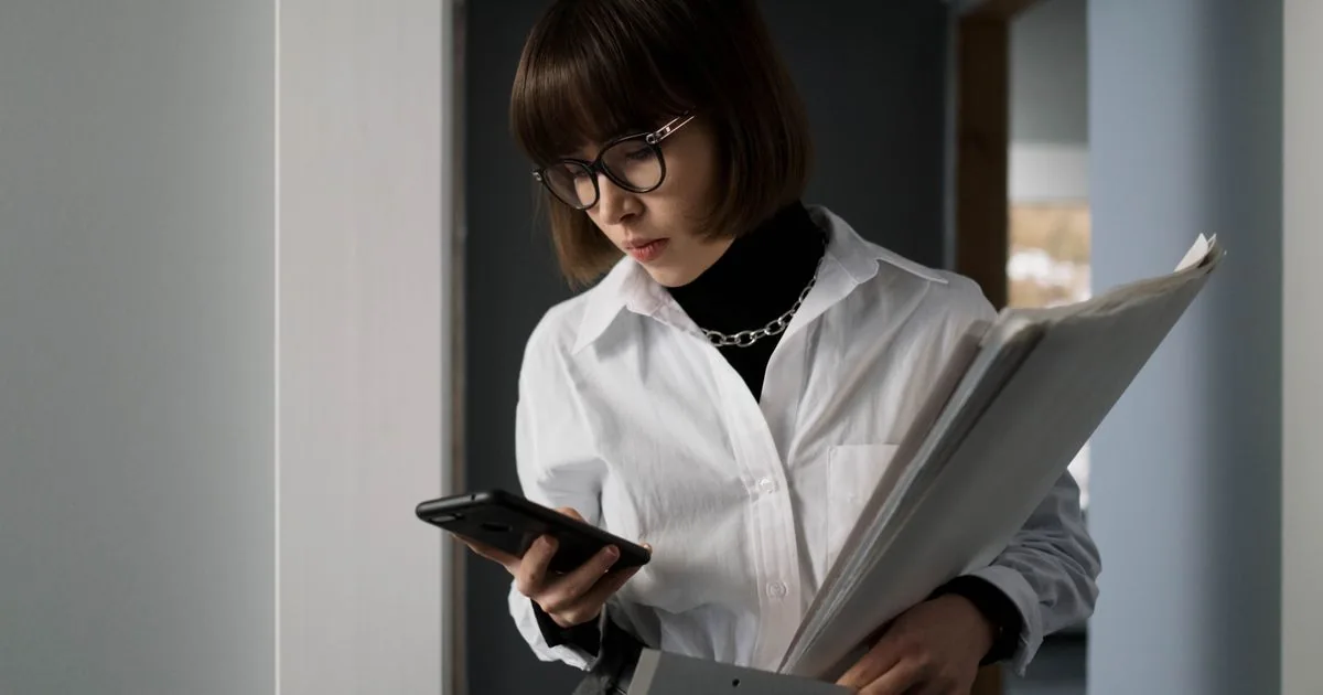 Mujer revisando una carpeta de documentos financieros mientras consulta su celular