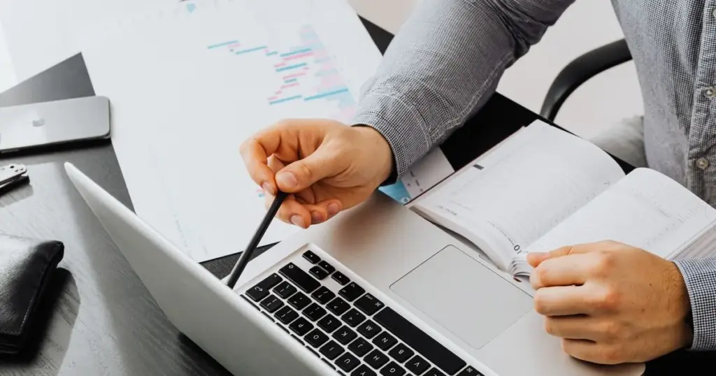 Persona comparando cifras en una laptop junto a gráficas impresas y una agenda