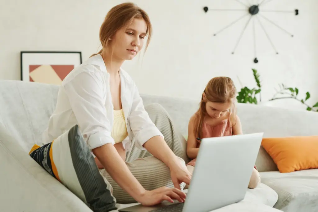 Madre soltera trabajando en su laptop junto a su hija en el sofá de casa