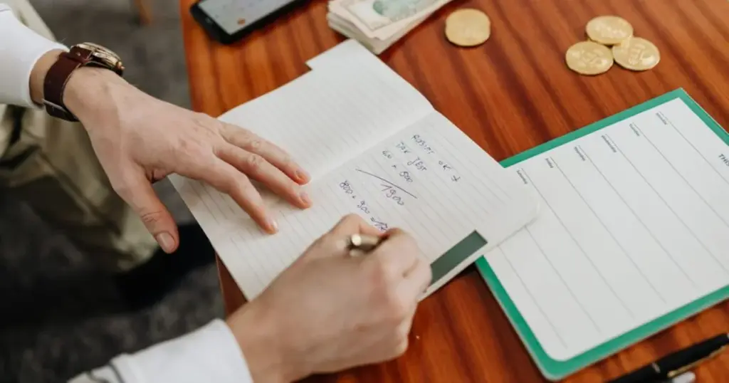 Persona escribiendo cálculos de presupuesto en un cuaderno junto a dinero en efectivo