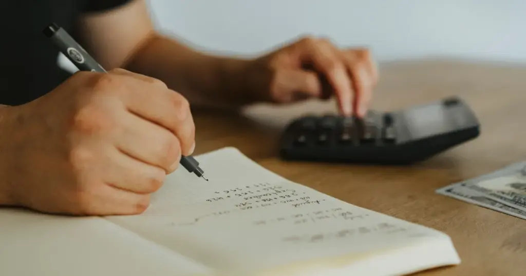 Manos escribiendo cálculos de presupuesto en cuaderno con calculadora